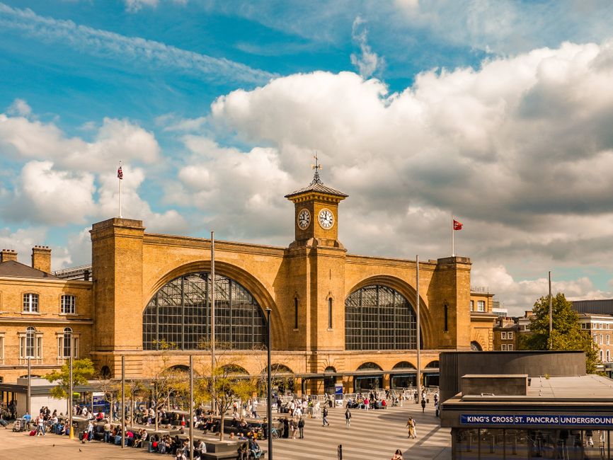 The image depicts King's Cross railway station in London, with its characteristic large arched windows and a central clock tower topped by a weather vane. In front of the station, there is a busy pave