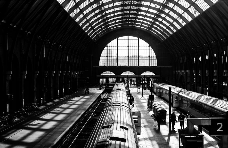 Inside a large train station with a high, arched glass ceiling allowing natural light to cast shadows on the platform. Multiple train carriages are visible, with one in the foreground being loaded or unloaded. Several people are present on the platform, some walking and others standing near the trains, possibly waiting or coordinating a home relocation or moving process. The station features structural beams supporting the glass roof, and a large window at the far end provides additional light. Some luggage or boxes may be seen near the waiting passengers, reflecting the ongoing logistics of packing and moving. The scene captures a busy moment involving the transportation of furniture and boxes, typical of professional removals or long-distance moving services such as those offered by Man with Van Kings Cross.
