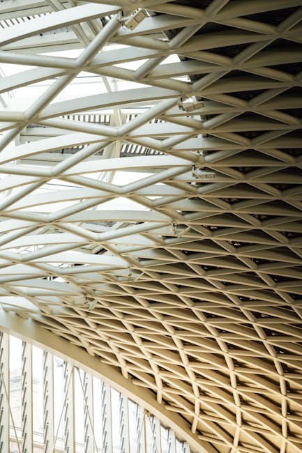 Close-up view of a large, modern architectural ceiling structure featuring intersecting white metal beams forming a geometric lattice pattern. The beams are supported by vertical metal columns visible at the lower left side of the image, and the design allows natural light to filter through the gaps. This structural detail is part of a contemporary building's roof, potentially in a public or commercial space, with a neutral colour palette and clean lines. Although not directly related to house removals or relocation, such architectural features are often present in residential or office buildings being prepared for moving or renovation. Man with Van Kings Cross may utilize similar structural visuals in their content to subtly convey the context of building or home relocation projects involving complex loading and transport logistics.