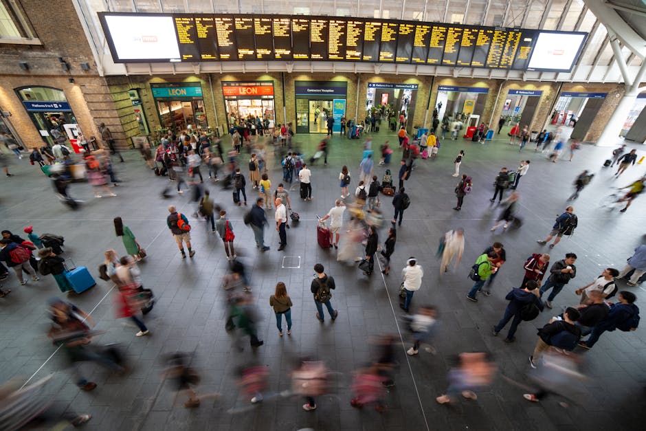 Inside a busy train station, a large crowd of travelers and commuters are moving in various directions across the open concourse. The scene shows numerous people walking, some with luggage, backpacks, or shopping bags, and a few using trolleys or carrying portable cases. The station has high ceilings with support beams, and an expansive digital departure board displaying train schedules hanging prominently overhead. Glass doors and ticketing counters are visible in the background, along with storefronts such as Leon. The floor is tiled, and lighting from above illuminates the area evenly. Some individuals are engaged in loading or unloading suitcases onto luggage trolleys, and others are waiting or walking towards platform entrances, reflecting the typical environment of home relocation or furniture transport preparations within a transport hub. Man with Van Kings Cross occasionally operates within such locations, assisting clients with packing and moving logistics as part of professional removals services.