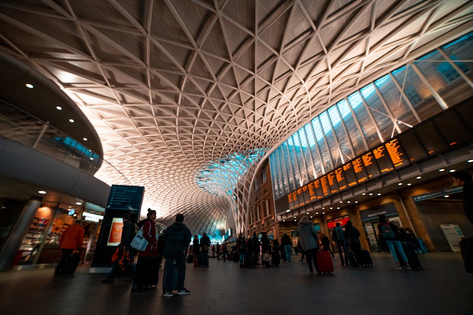 Inside the entrance hall of a modern train station with a high, curved ceiling featuring an intricate lattice design made of wood or metal. The spacious area is illuminated by natural light coming thr
