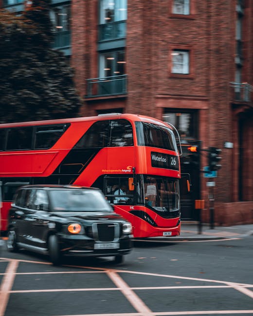 A red double-decker bus displaying route number 26 and destination Waterloo is passing through a city street with tall red brick residential buildings featuring large windows and balconies on either side. A black classic-style taxi is driving nearby, slightly blurred to indicate motion. The street is marked with yellow lane lines, and traffic lights are visible at the intersection. The scene is captured during daylight hours with overcast lighting. This setting relates to urban transportation and movement logistics, relevant to house removals and moving services provided by Man with Van Kings Cross, as shown in the webpage about loading bay and street closure rules in Kings Cross (Camden).
