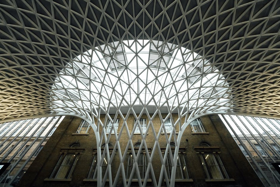 A view from inside a building showcasing a modern architectural structure with an intricate, lattice-like white roof covering a circular opening, allowing natural light to filter through. Below the ceiling, an older brick building with multiple windows is visible, blending contemporary design with historic architecture. The view emphasizes the spacious interior, highlighting the contrast between the sleek, geometric ceiling pattern and the traditional brick facade outside. This setting is part of the loading bay area at Kings Cross, where furniture and packing materials may be prepared for home relocation, with visible support structures such as trolleys or loading equipment occasionally used by Man with Van Kings Cross during the furniture transport and packing process involved in house removals and street closure arrangements.