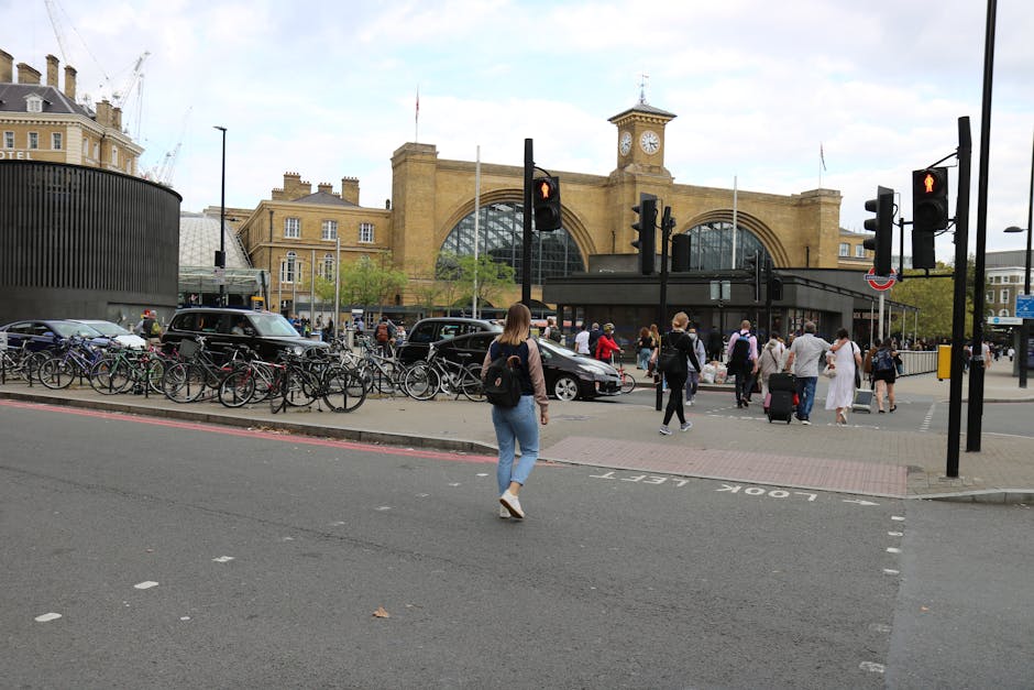 A busy street scene outside King's Cross Station in London during daytime, featuring a diverse group of pedestrians crossing the road and walking along the pavement. In the foreground, a woman with brown hair, wearing a dark jacket and light blue jeans, is walking towards the station entrance, which is visible behind a row of bicycles parked on a bike rack. Several other pedestrians, some with luggage or bags, are heading in various directions near the station's large brick facade with arched windows and a clock tower. To the left, parked cars and a black van are aligned along the curb, with a section of loading area marked on the street. The surrounding environment includes urban infrastructure like traffic lights, street signs, and black bollards, highlighting an active transportation hub. This scene is typical of home relocation or moving activities in the area, supported by [COMPANY_NAME] specialized in removals and furniture transport, emphasizing the logistical aspect of moving logistics in Central London.