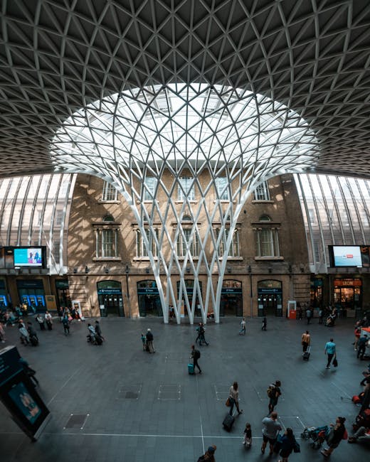 A busy street scene outside King's Cross Station in London during daytime, featuring a diverse group of pedestrians crossing the road and walking along the pavement. In the foreground, a woman with brown hair, wearing a dark jacket and light blue jeans, is walking towards the station entrance, which is visible behind a row of bicycles parked on a bike rack. Several other pedestrians, some with luggage or bags, are heading in various directions near the station's large brick facade with arched windows and a clock tower. To the left, parked cars and a black van are aligned along the curb, with a section of loading area marked on the street. The surrounding environment includes urban infrastructure like traffic lights, street signs, and black bollards, highlighting an active transportation hub. This scene is typical of home relocation or moving activities in the area, supported by [COMPANY_NAME] specialized in removals and furniture transport, emphasizing the logistical aspect of moving logistics in Central London.