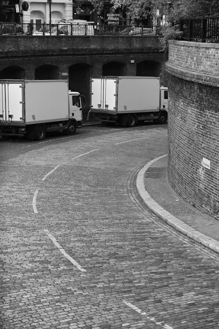 A black-and-white photograph showing a narrow, curved cobblestone street in an urban area, likely part of a city centre, with two large white commercial boxes or containers mounted on truck chassis parked along the street. The trucks are positioned alongside a brick wall on the right side of the image, which supports a curved sidewalk and a metal fence at the top with decorative railing. In the background, there is a stone structure with multiple arches, possibly a bridge or viaduct, and greenery including trees is visible beyond the fence. The scene is evenly lit, capturing the loading process involved in a house removal or home relocation, with the trucks ready for furniture and boxes. The environment suggests a typical urban setting where [COMPANY_NAME], such as Man with Van Kings Cross, would facilitate moving services, including furniture transport and packing during the loading phase of an urgent same-day move in Kings Cross.