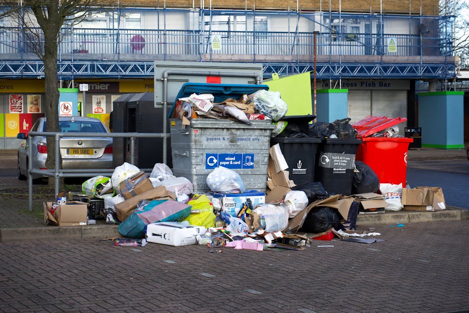 A collection of overflowing rubbish and recycling bins situated on a paved pavement in Kings Cross, with various waste items including cardboard boxes, plastic bags, paper, and household packaging spilling onto the ground. The bins are positioned next to a metal railing, with some cardboard boxes placed on the pavement nearby. Behind the waste collection area, there is a building with a blue scaffolding structure and signage for local businesses, as well as a parked silver car with a visible UK registration plate. The scene is lit with natural daylight, indicating daytime, and captures the typical environment where household waste is temporarily stored before disposal or collection, seen in the context of home relocation and moving logistics handled professionally by Man with Van Kings Cross.