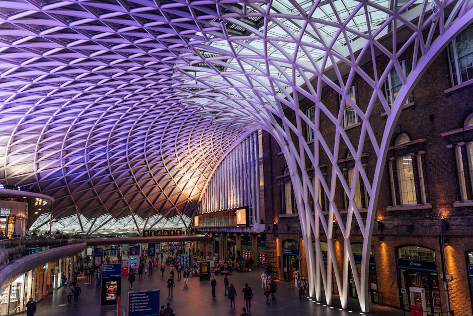 A spacious indoor area of King's Cross railway station featuring a modern, intricate lattice ceiling structure illuminated with purple and white lighting. The high, curved ceiling spans across the station, supported by large white beams forming a web-like pattern. Below, there are ticketing and information displays, digital screens, and a variety of travelers walking, some carrying luggage or boxes in a house removals context. The station's brick walls and large windows are visible to the right, contrasting with the contemporary ceiling design. The environment is lively with groups of people moving through the space, and the lighting highlights the architectural details. This indoor setting illustrates the flow of people during home relocation or furniture transport activities, where clear pathways and organized areas facilitate efficient packing and moving processes, supported by services like Man with Van Kings Cross.