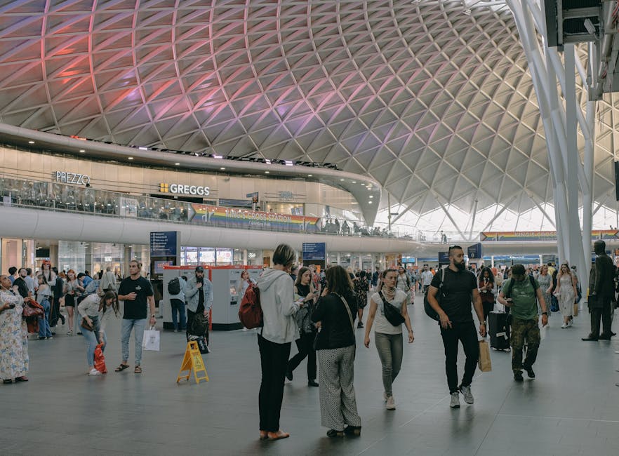 Inside a large modern train station with a high, geometrically patterned ceiling illuminated by soft pink and purple lighting. The spacious area is filled with numerous travelers walking, waiting, and carrying luggage or shopping bags. Several people are engaged in conversations or looking at their phones. In the background, there are digital information screens and a cafe area with visible signs for Prezzo and Greggs. The station's open-plan design includes a mezzanine level with additional shoppers and seating. The scene captures the busy atmosphere typical of home relocation logistics, with individuals preparing for their journeys or waiting to load luggage into waiting vehicles. Outdoors, a vehicle is being loaded with cardboard boxes and packed furniture covered in protective blankets, indicating a professional furniture transport and removal process. The company Man with Van Kings Cross is involved in the loading process, assisting with packing and moving services in preparation for local or long-distance house removals.